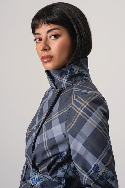 A woman with a dark hair is shown in profile wearing RainSisters high-neck jacket Dutchess in Blue with a blue, gray, and white plaid pattern and subtle floral accents near the hem, against a plain light gray studio background.