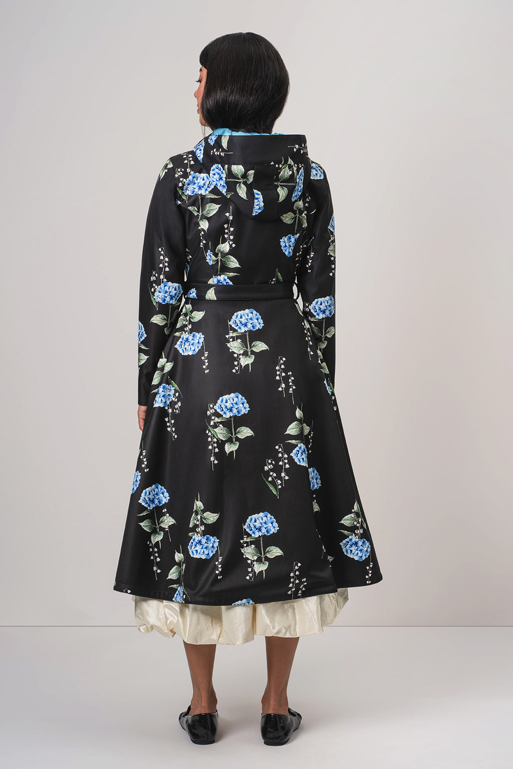 A woman viewed from the back wearing RainSisters black long-sleeve coat Flower Moon with a blue and green floral print, layered over a cream ruffled underskirt and black flats, against a light gray studio background.