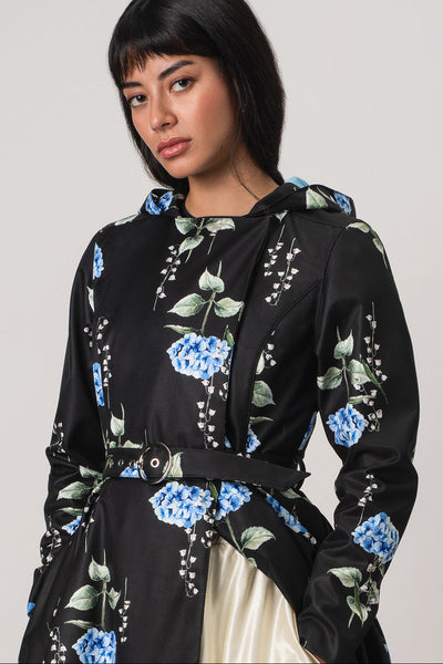 A woman wearing RainSisters black hooded raincoat Flower Moon with a blue and green floral print, styled with a matching belt and a cream satin skirt, against a plain light gray background.