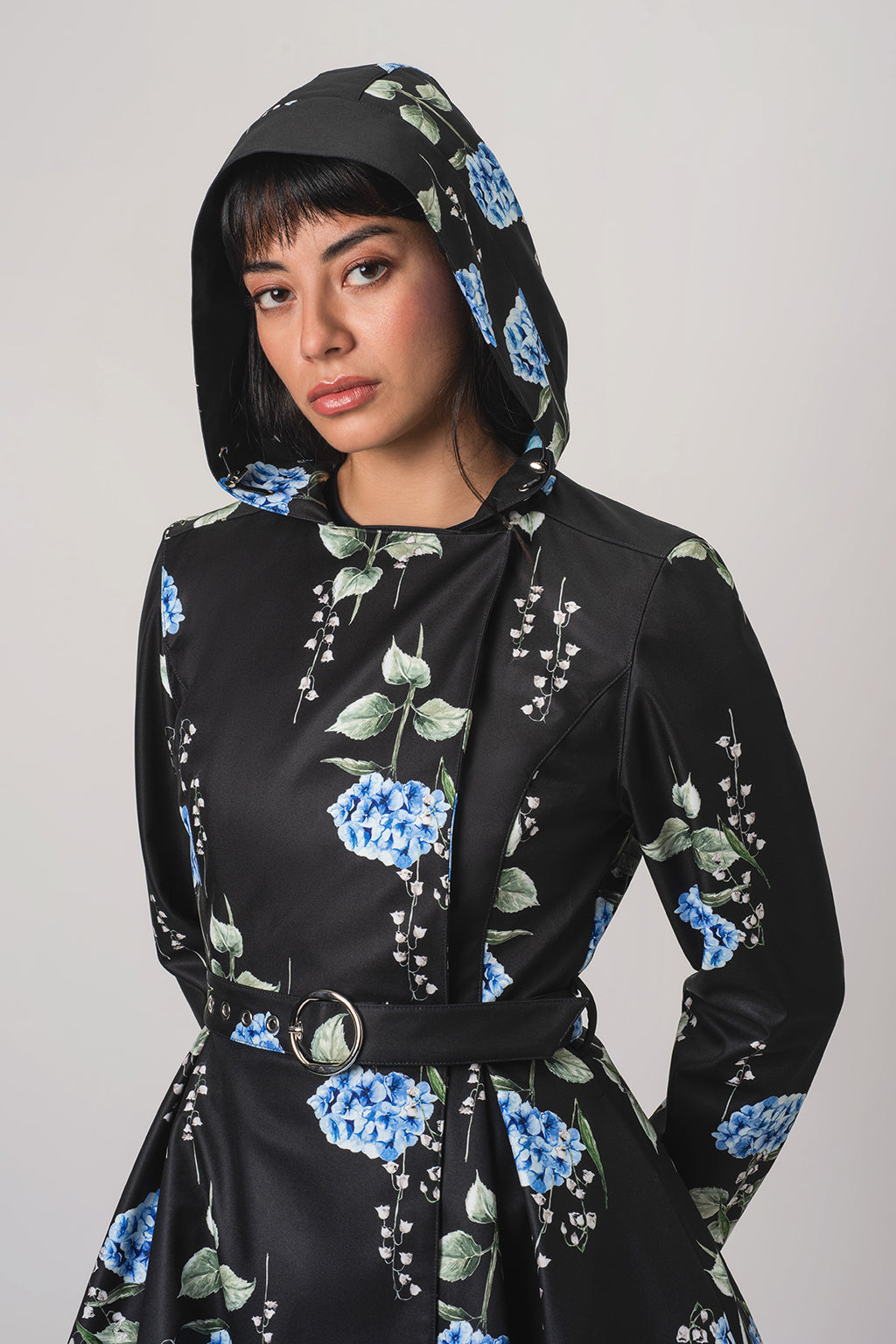 A woman wearing RainSisters black hooded raincoat Flower Moon with a blue and green floral print and hood, styled with a matching belt and a cream satin skirt, against a plain light gray background.