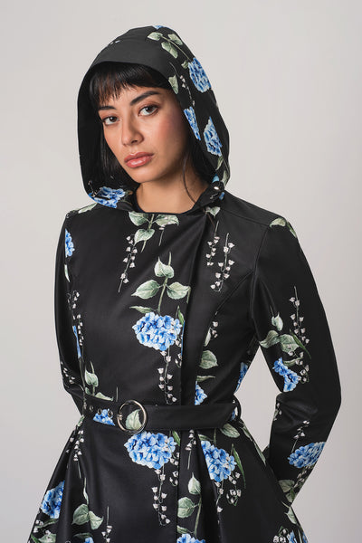 A woman wearing RainSisters black hooded raincoat Flower Moon with a blue and green floral print and hood, styled with a matching belt and a cream satin skirt, against a plain light gray background.
