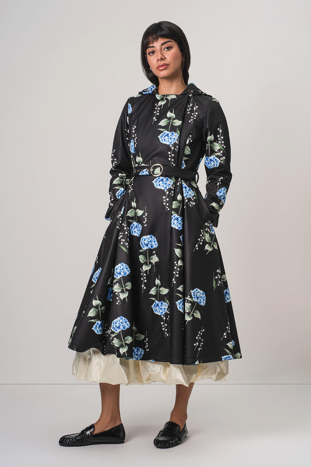 A woman modeling RainSisters black hooded long-sleeve coat Flower Moon with a blue and green floral print, featuring a matching belt and a cream ruffled underskirt, paired with black loafers against a light gray studio background.