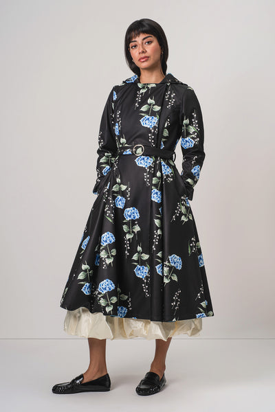 A woman modeling RainSisters black hooded long-sleeve coat Flower Moon with a blue and green floral print, featuring a matching belt and a cream ruffled underskirt, paired with black loafers against a light gray studio background.