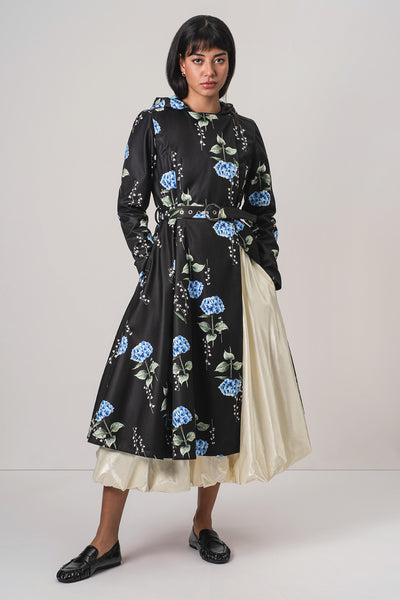 A model wearing RainSisters black long-sleeve midi raincoat Flower Moon with a blue floral print, a matching black belt, and a layered cream satin underskirt, paired with black loafers against a light gray studio background.