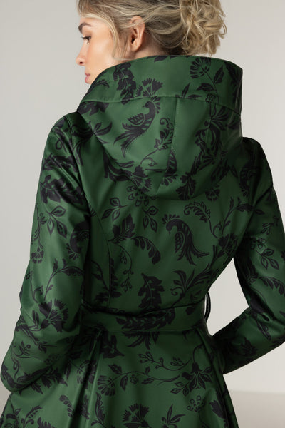 A woman shown from the back wearing RainSisters dark green belted coat Forest Flower with black damask-style floral and bird pattern, photographed against a plain light gray studio background.