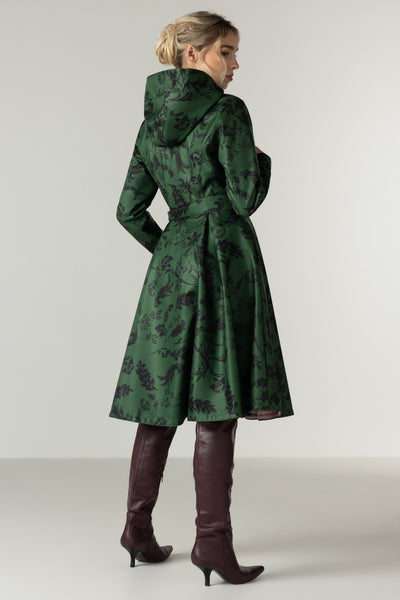 A woman modeling RainSisters green hooded coat Forest Flower with a dark floral jacquard pattern, shown from the back and three-quarter side view against a neutral studio background.