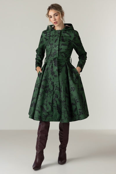 A woman modeling RainSisters dark green hooded coat Forest Flower with a black floral pattern, paired with dark brown over-the-knee boots, against a neutral light gray studio background.