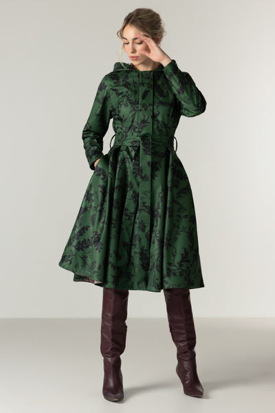 A woman models RainSisters dark green hooded waterproof coat Forest Flower with a black floral pattern, paired with dark brown knee-high boots, against a neutral light gray studio background.