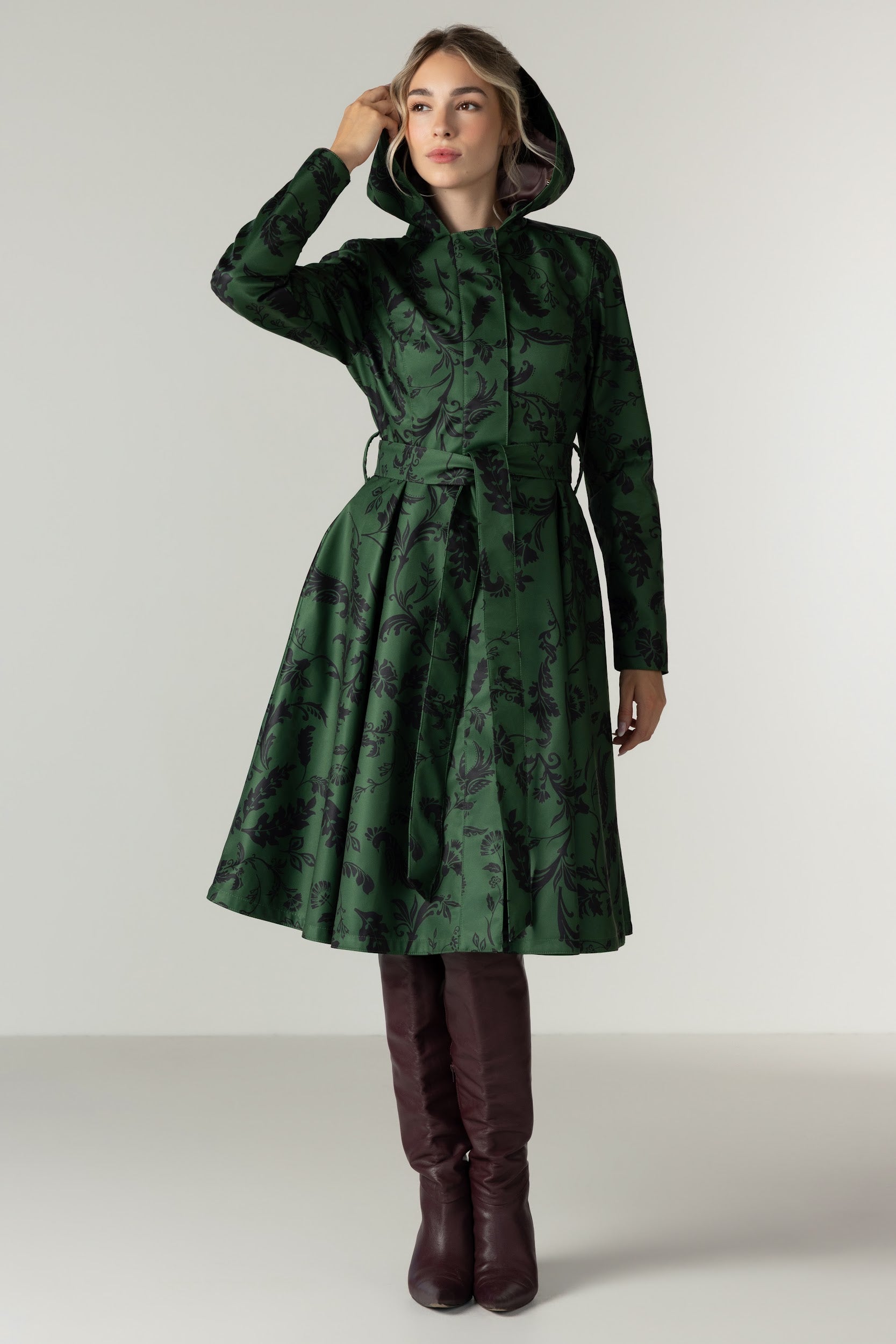 A woman models RainSisters dark green hooded waterproof coat Forest Flower with a black floral pattern, featuring a tie waist belt and long sleeves, paired with dark brown knee-high boots against a neutral studio background.