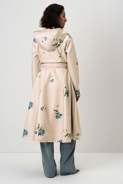 A woman shown from the back wearing RainSisters light beige hooded raincoat Gentle Bloom with a blue and green floral print, paired with wide-leg blue jeans, against a plain light gray studio background.