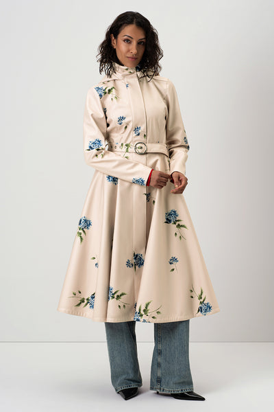 A model wears RainSisters high-neck, long-sleeved cream-colored coat Gentle Bloom with blue floral print and a matching belt, styled with wide-leg blue jeans and black pointed shoes against a light gray studio background.