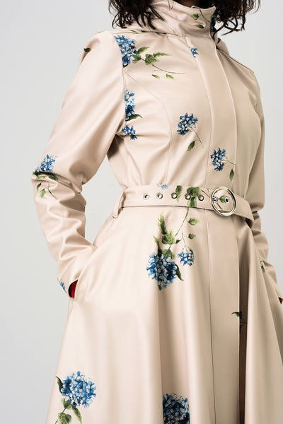 A close-up of RainSisters beige trench coat Gentle Bloom with blue floral prints and a matching belt with a silver buckle, shown on a model against a light gray background.