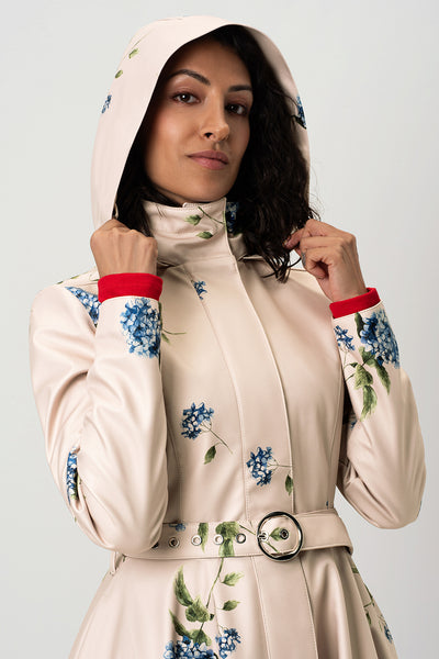 A close-up of RainSisters beige trench coat Gentle Bloom with blue floral prints and a matching belt with a silver buckle and hood on, shown on a model against a light gray background.