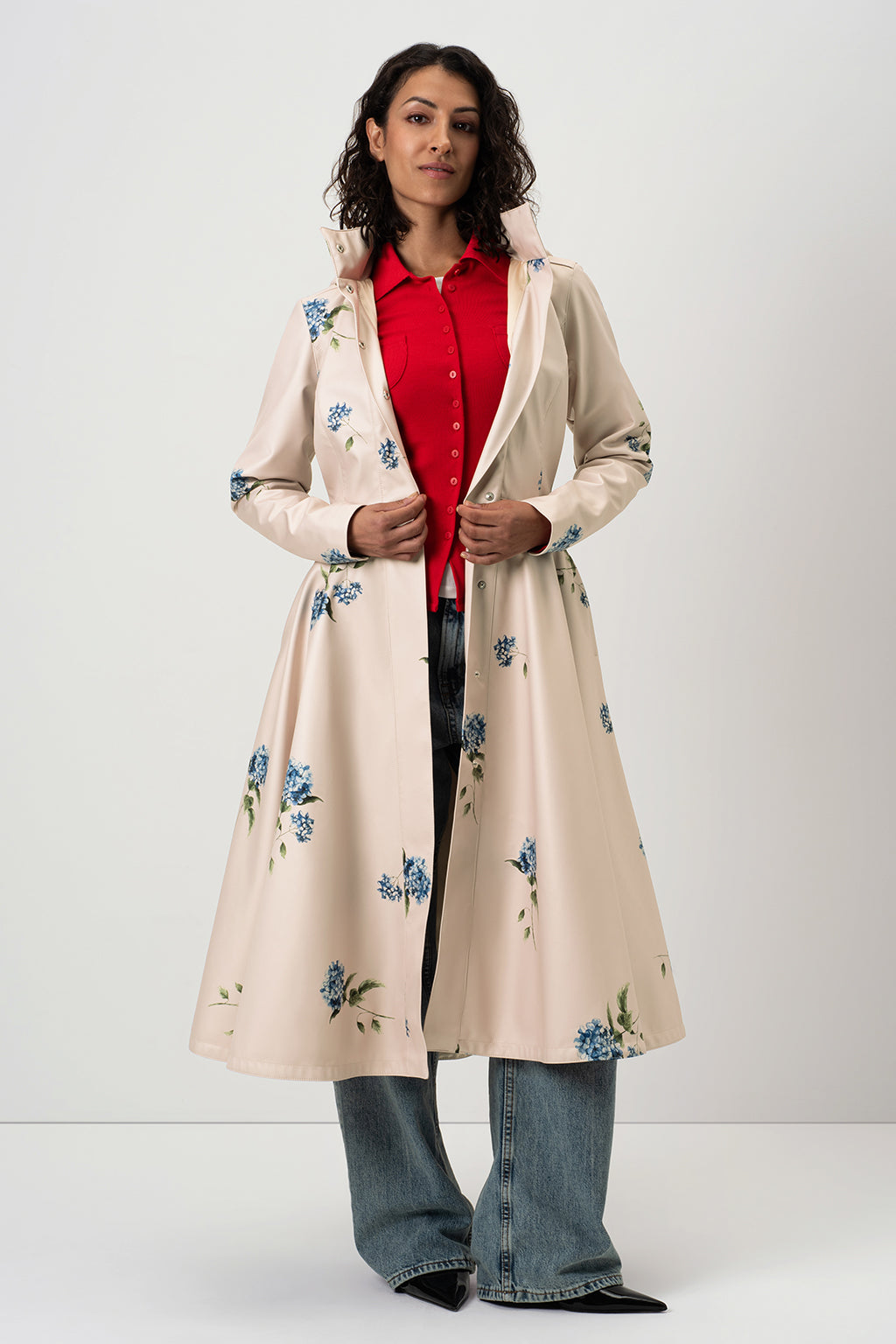 A woman models RainSisters long cream-colored hooded trench coat Gentle Bloom with blue floral prints, worn over a red button-up shirt and blue jeans against a plain light gray studio background.