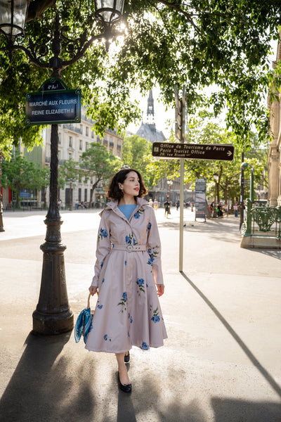 A woman wearing RainSisters light pink floral trench coat Gentle Bloom and blue top walks along a sunlit city street lined with trees and lampposts.