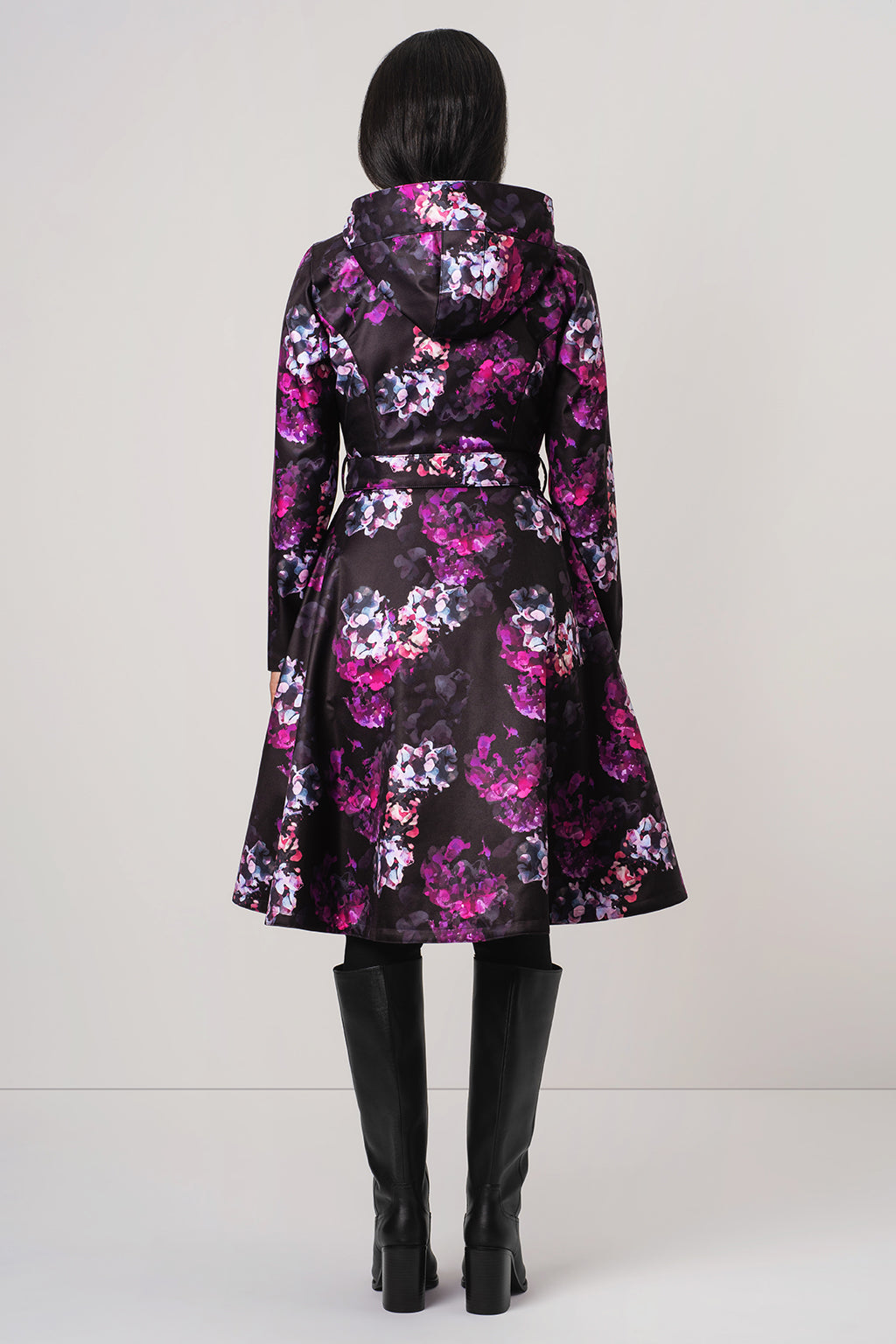A model viewed from the back wearing RainSisters black hooded coat Hortense with a vibrant pink, purple, and white floral pattern, paired with black knee-high boots against a light gray studio background.