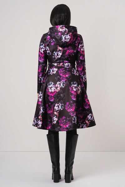 A model viewed from the back wearing RainSisters black hooded coat Hortense with a vibrant pink, purple, and white floral pattern, paired with black knee-high boots against a light gray studio background.