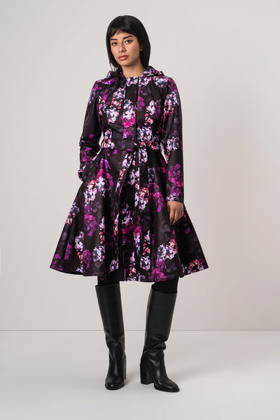 A woman modeling RainSisters black hooded raincoat Hortense with a vibrant pink, purple, and white floral pattern, styled with black knee-high boots against a light gray studio background.