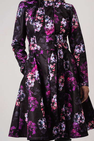 RainSisters long-sleeve black satin-like raincoat Hortense with a vibrant purple, pink, and light floral print, featuring a tie waist belt and a flared skirt.
