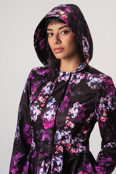 A woman wearing RainSisters hooded raincoat Hortense with a purple, pink, and white floral pattern on a black background, photographed against a plain light gray studio backdrop.