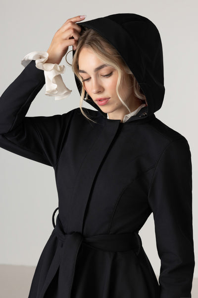 A woman wearing RainSisters black hooded waterproof coat Midnight Black with a matching waist tie, styled with a white ruffled collar detail, set against a plain light gray background.