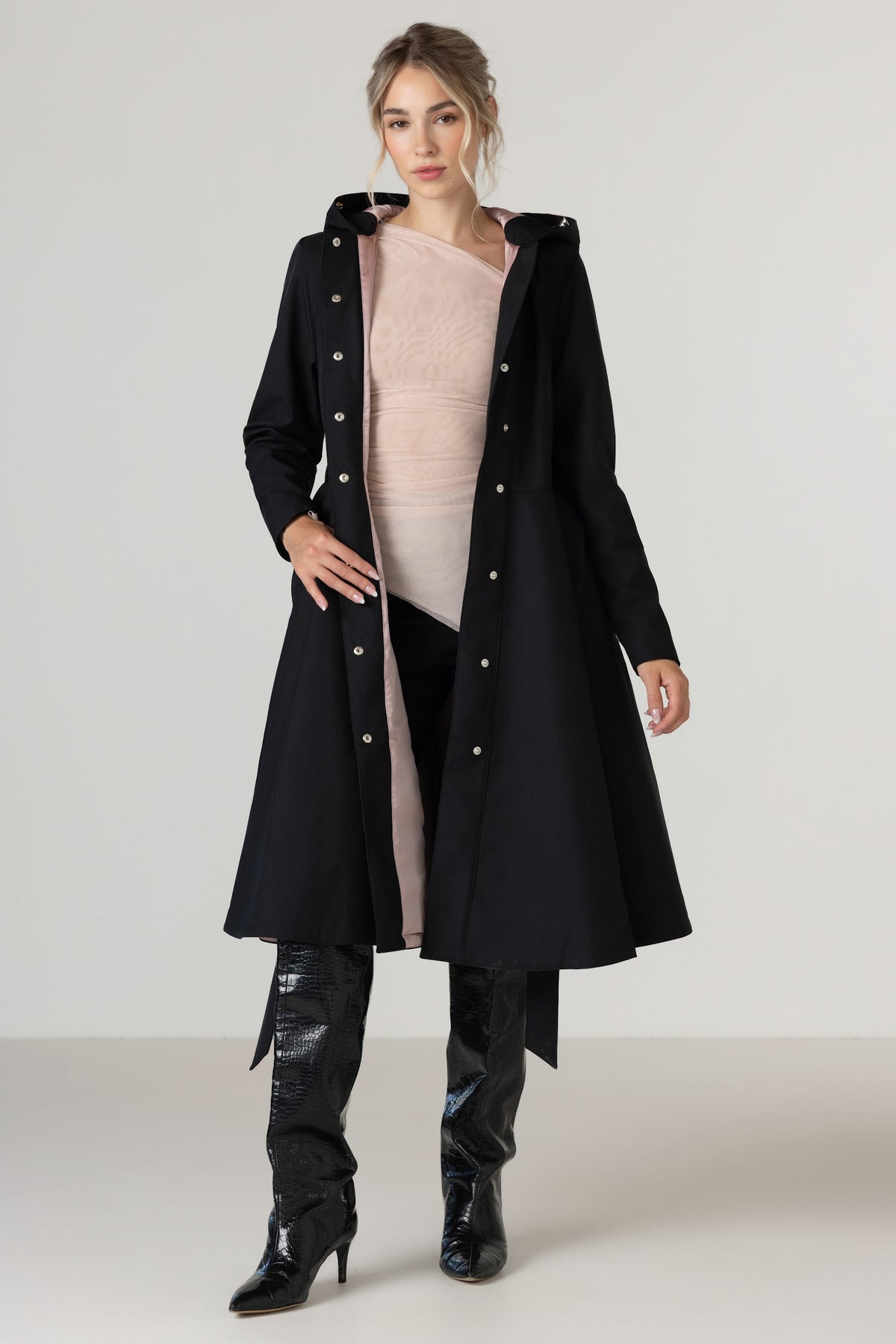 A woman models RainSisters long black hooded trench coat Midnight Black over a light pink top, black pants, and black knee-high boots against a plain light gray studio background.