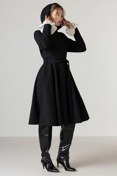 A model wearing RainSisters black long-sleeve raincoat Midnight Black with a hood, styled with ruffled blouse, and shiny black patent leather boots, standing against a light gray studio background.
