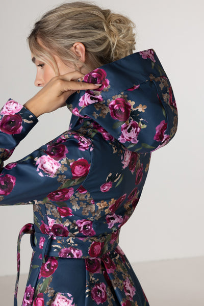 A woman wearing RainSisters navy blue  floral waterproof coat Misty Rose with pink and purple roses, shown from the back and side against a plain light background.