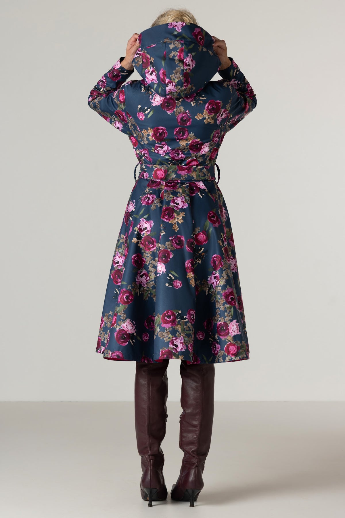 A woman viewed from behind wearing RainSisters dark denim blue floral coat Misty Rose with pink and purple flowers, paired with dark brown knee-high boots, against a light gray studio background.