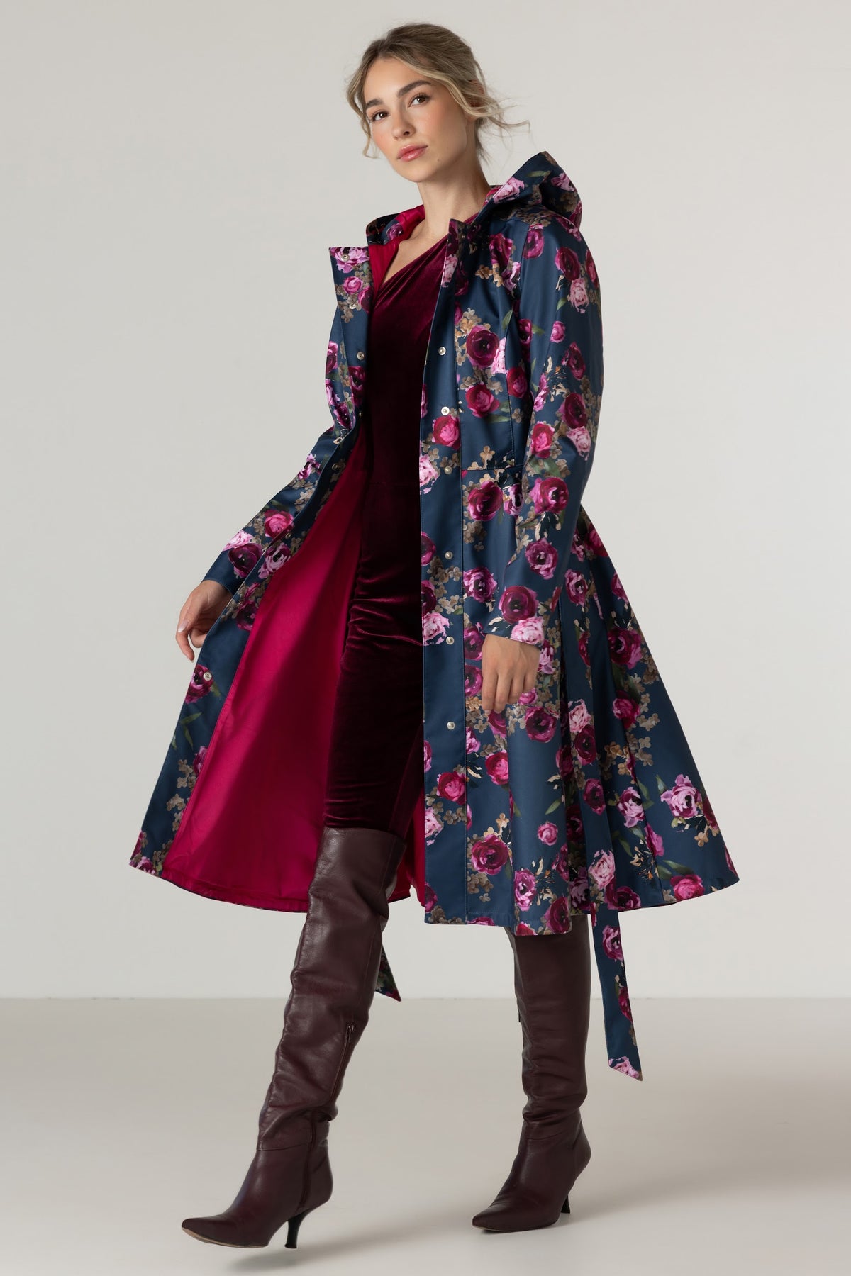 A woman models RainSisters long navy blue hooded raincoat Misty Rose with a vibrant pink and purple floral print and bright pink inner lining, worn over a deep burgundy velvet dress and paired with dark brown knee-high leather boots, set against a plain light gray studio background.