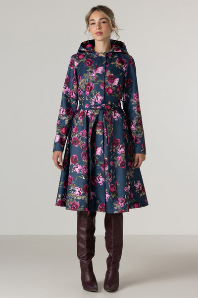 A woman models RainSisters dark navy blue hooded raincoat Misty Rose with a vibrant pink and purple floral print, styled with dark brown knee-high leather boots against a neutral light gray studio background.