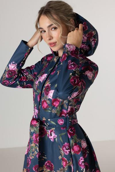 A woman wearing RainSisters navy blue hooded waterproof coat Misty Rose with a pink and purple floral print, posing against a light gray studio background.