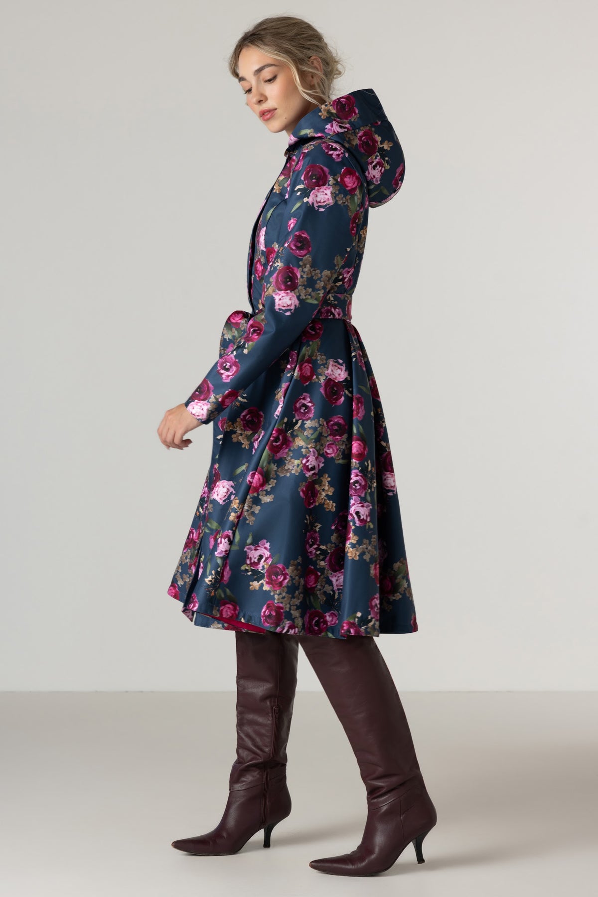 A woman models RainSisters navy blue hooded raincoat Misty Rose with a vibrant pink, red, and purple floral pattern, paired with dark brown knee-high leather boots against a neutral studio background.