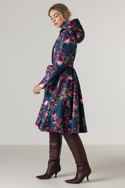 A woman models RainSisters navy blue hooded raincoat Misty Rose with a vibrant pink, red, and purple floral pattern, paired with dark brown knee-high leather boots against a neutral studio background.