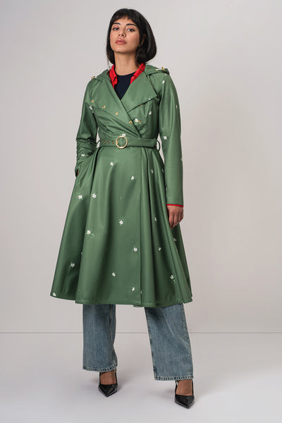 A woman models RainSisters green double breasted trench coat Primrose in Bloom with white floral print, a matching belt with a gold buckle, styled with blue jeans and black heels against a light gray studio background.