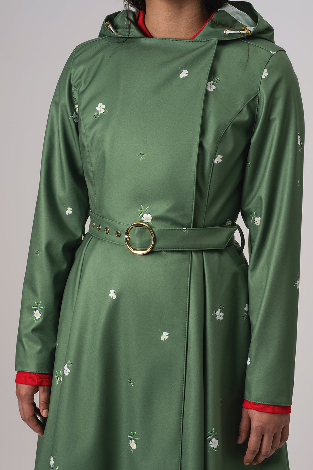 RainSisters green hooded raincoat Primrose in Bloom with a matching waist belt and gold-tone ring buckle, decorated with small white floral prints. The coat is shown on a model against a neutral gray background.