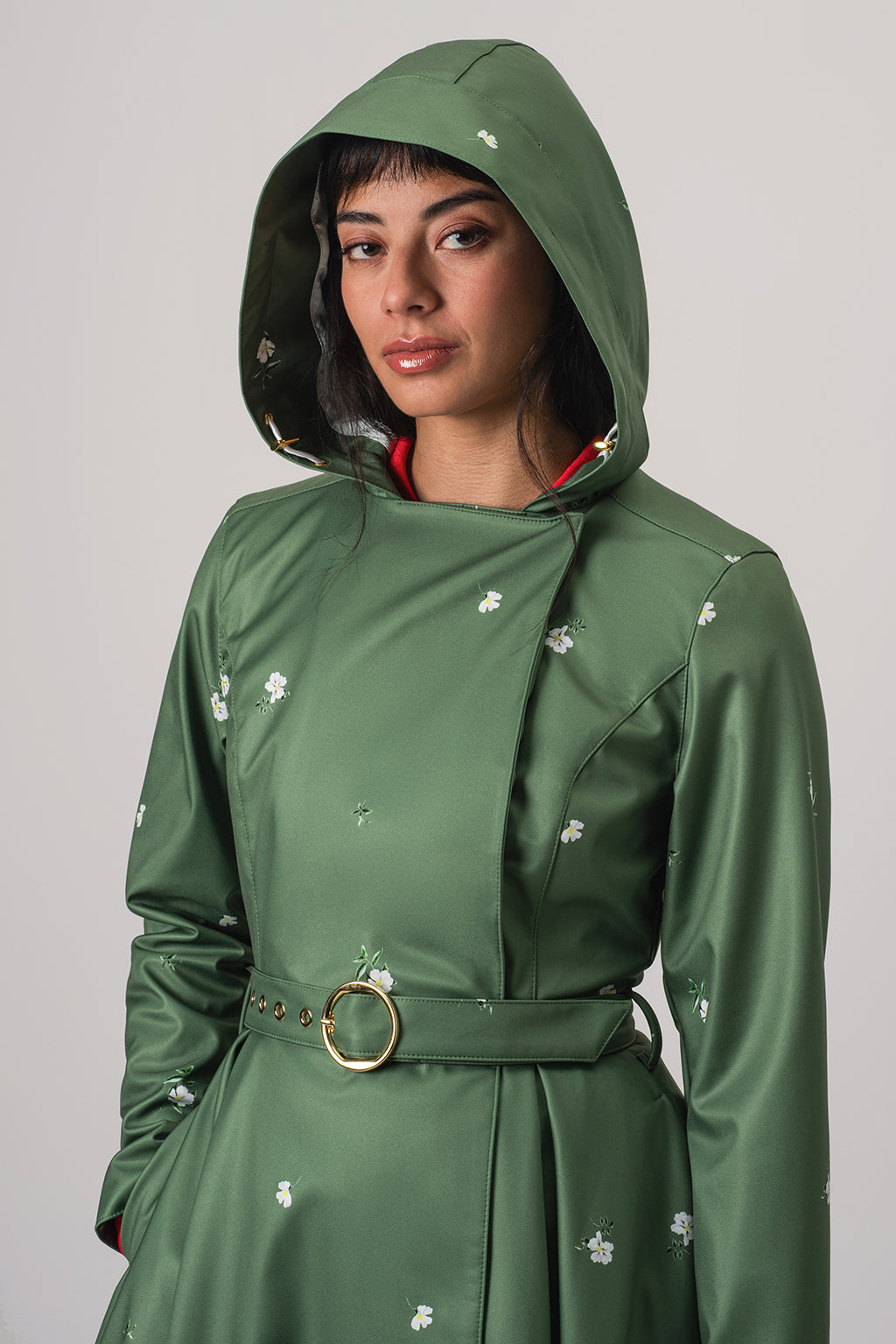 A woman wearing RainSisters green hooded raincoat Primrose in Bloom with small white floral print and a matching belt with a gold ring, photographed against a plain light gray studio background.
