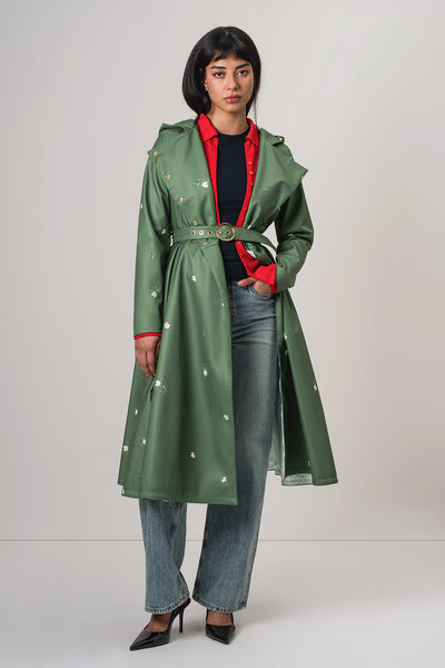 A model wearing RainSisters green belted trench coat Primrose in Bloom with small white floral prints, layered over a red collared shirt, black top, blue jeans, and black heels, photographed against a light gray studio background.