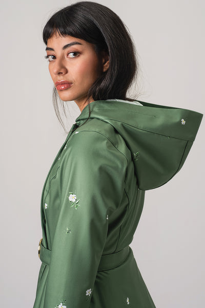 A woman wearing RainSisters sage green hooded raincoat Primrose in Bloom with white floral details, shown in a side profile against a light gray studio background.