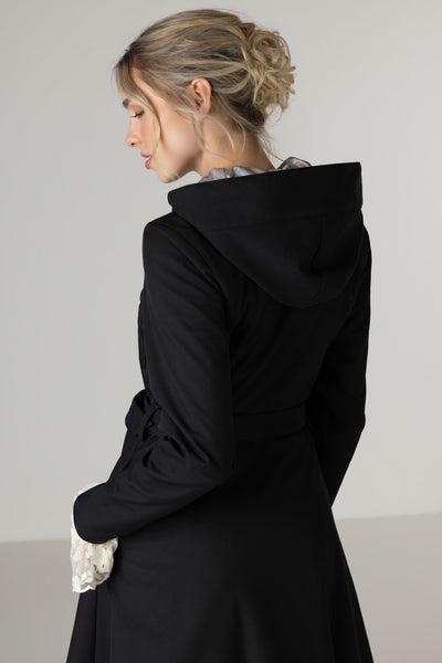 A woman shown from the back wearing RainSisters black hooded raincoat Queen of Spades against a plain light gray studio background.