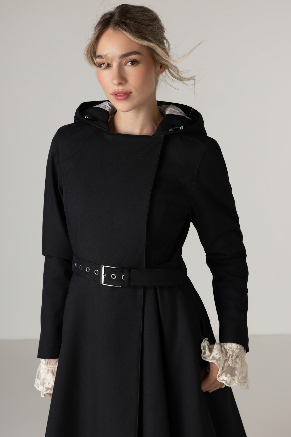 A woman wearing RainSisters black hooded longline trench coat Queen of Spades with a matching belt styled with lace ruffled cuffs, photographed against a plain light gray studio background.