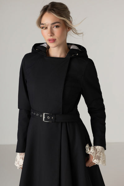 A woman wearing RainSisters black hooded longline trench coat Queen of Spades with a matching belt styled with lace ruffled cuffs, photographed against a plain light gray studio background.