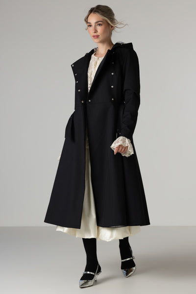 A model wearing RainSisters waterproof long black double-breasted coat Queen of Spades over a cream-colored dress, paired with black tights and silver pointed-toe heels, photographed against a neutral light gray studio background.