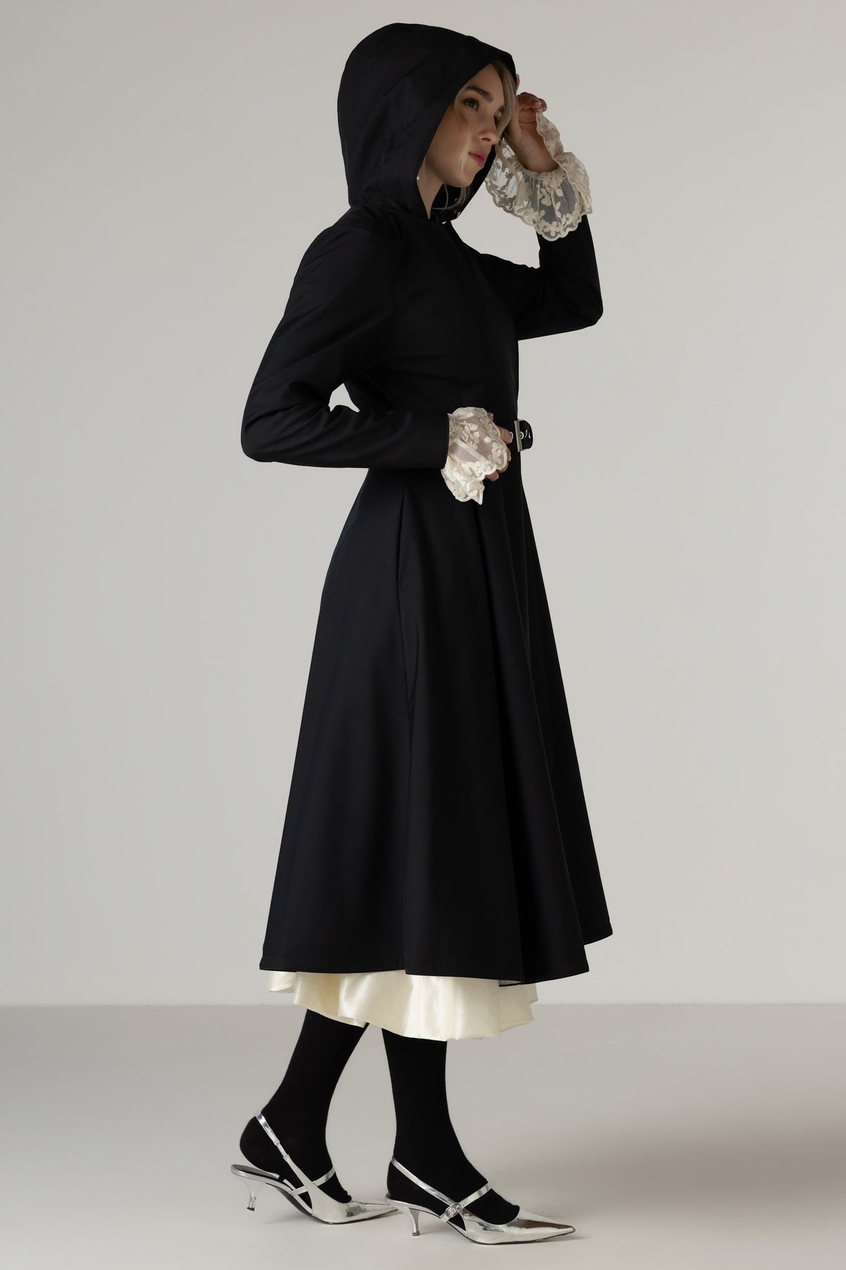 A model is shown from the side wearing RainSisters black hooded long-sleeve trench coat Queen of Spades with a cream underskirt, black tights, and black-and-white kitten heels against a neutral studio background.