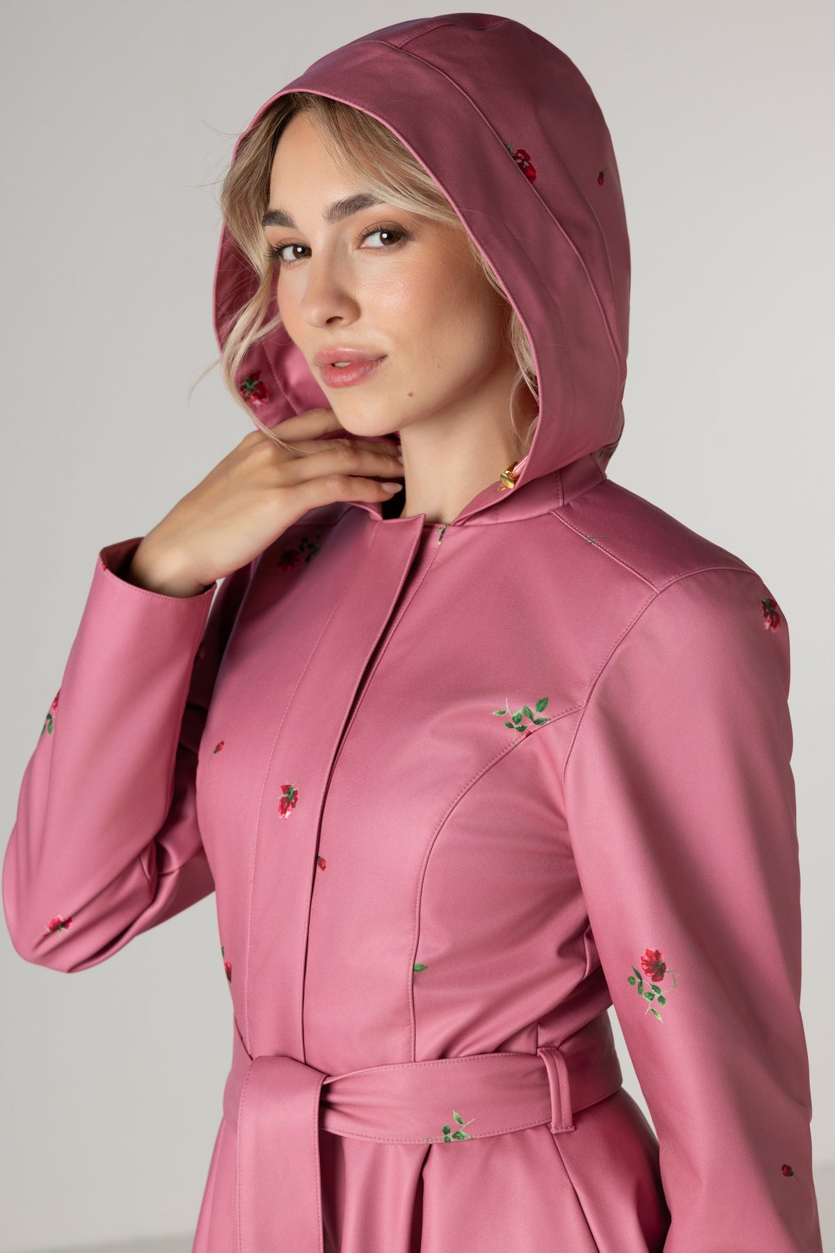 A woman wearing RainSisters dusty pink hooded raincoat Raspberry Sorbet with a matching waist tie and small red rose flower print, photographed against a plain light gray studio background.