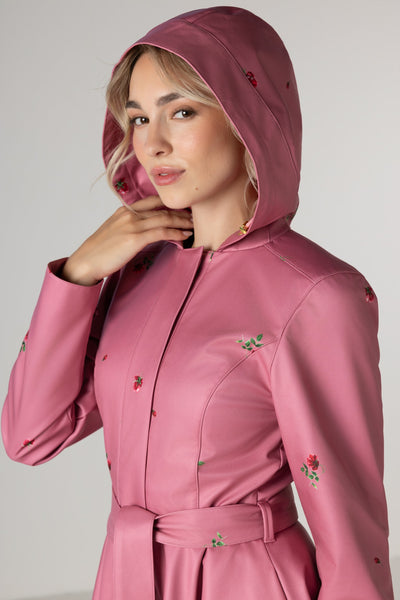 A woman wearing RainSisters dusty pink hooded raincoat Raspberry Sorbet with a matching waist tie and small red rose flower print, photographed against a plain light gray studio background.