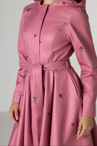 A woman wearing RainSisters pink hooded raincoat Raspberry sorbet with a matching waist tie and small red rose print, shown against a light gray studio background.