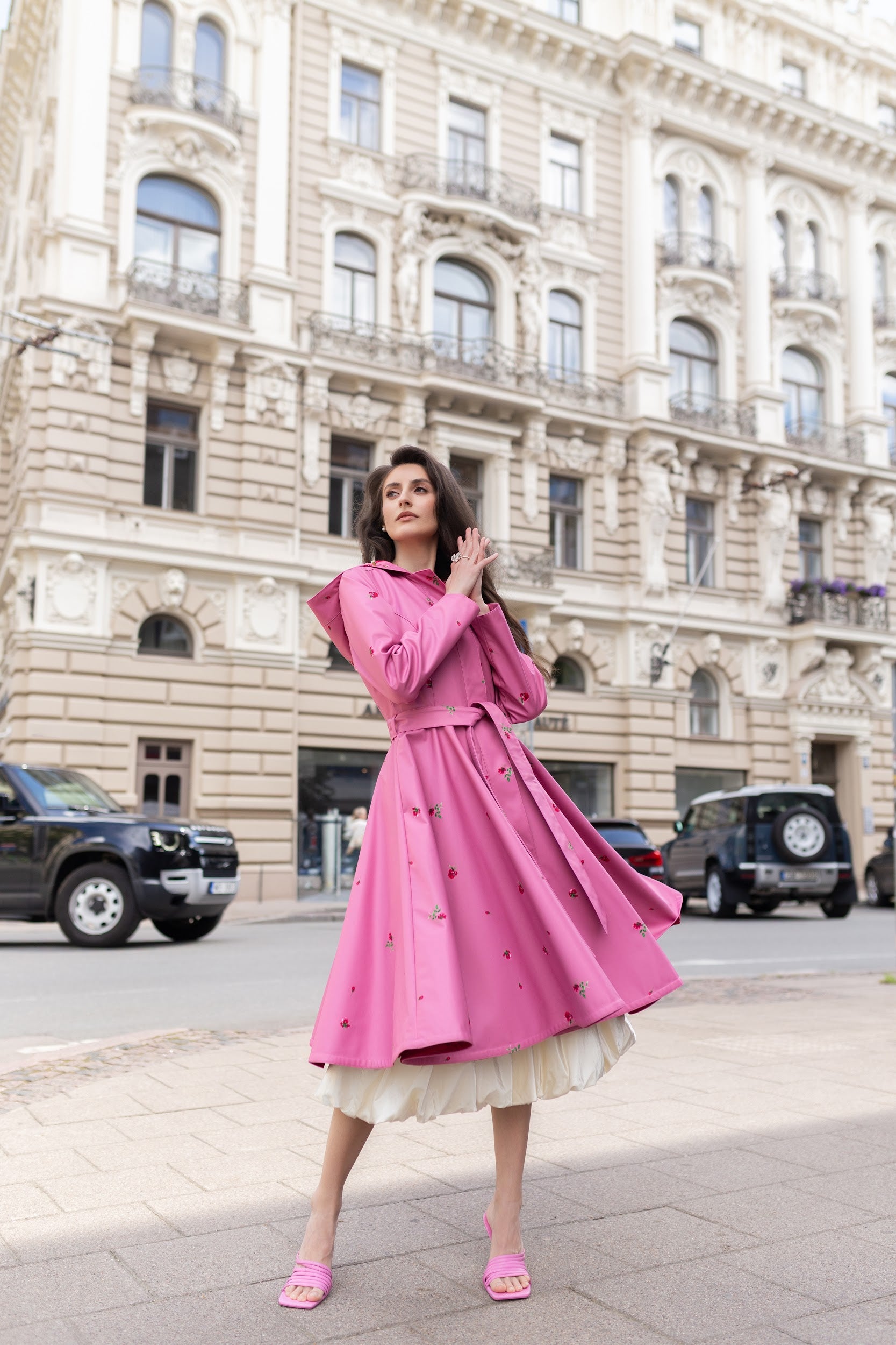 A woman stands on a city sidewalk in front of a large beige historic building, wearing RainSisters bright pink long-sleeve raincoat Raspberry Sorbet with a ruffled skirt underneath and pink pointed heels.