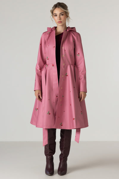 A woman stands against a plain light gray studio background wearing RainSisters long pink hooded raincoat Raspberry Sorbet with small multicolor rose print, a dark top, and dark brown knee-high boots.
