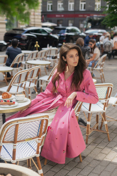 A woman wearing RainSisters pink waterproof fit and flared coat Raspberry Sorbet with small red floral print sits at an outdoor cafe table surrounded by woven chairs.